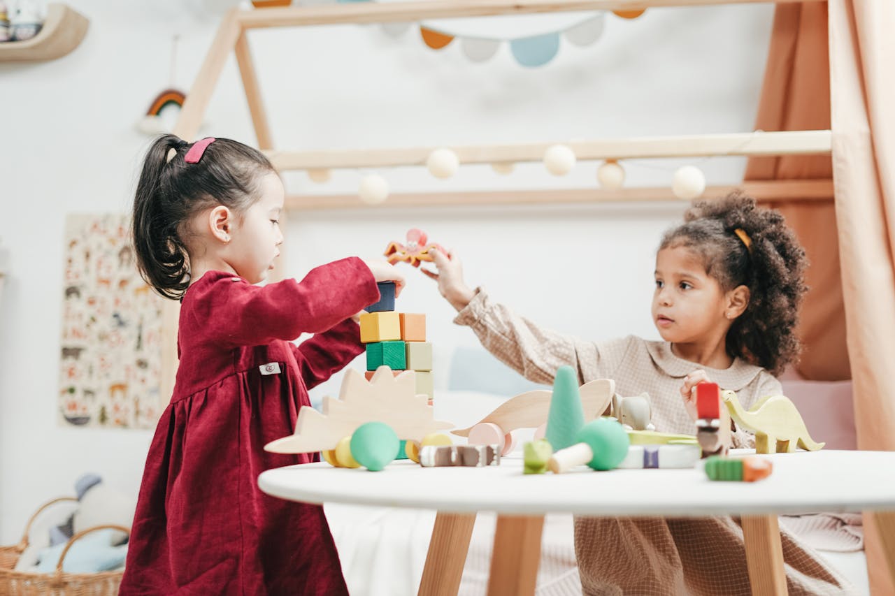 about-03 Two young girls enjoying playtime with wooden toys indoors in a warm, colorful playroom.